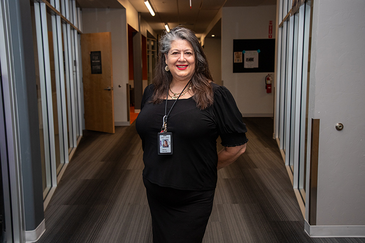 A woman with long gray hair wearing a black outfit smiles in a school hallway
