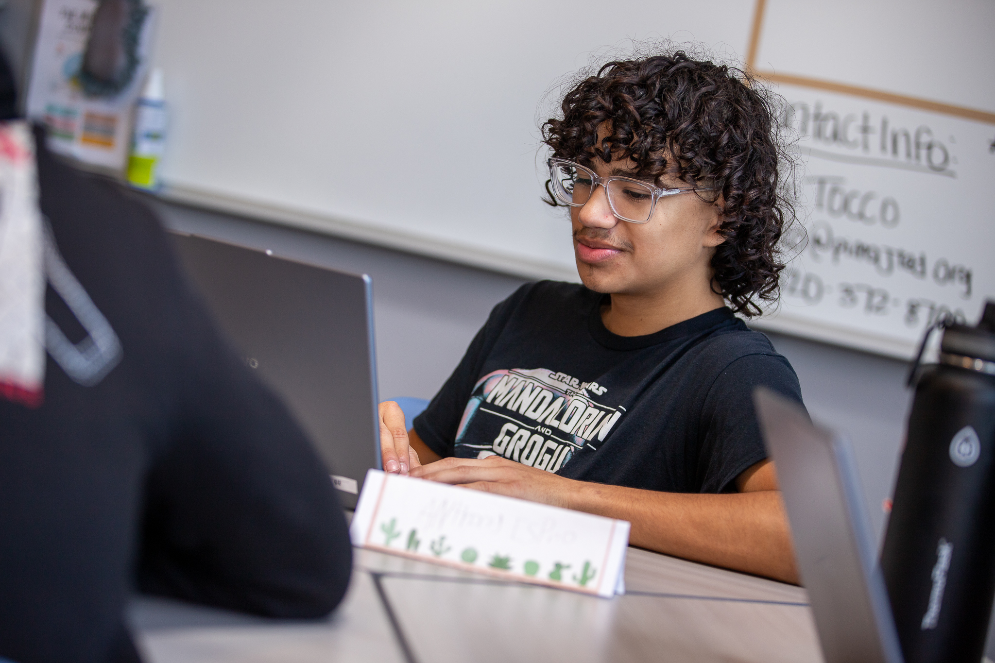 A teen boy in glasses smiles as he works on his laptop in class