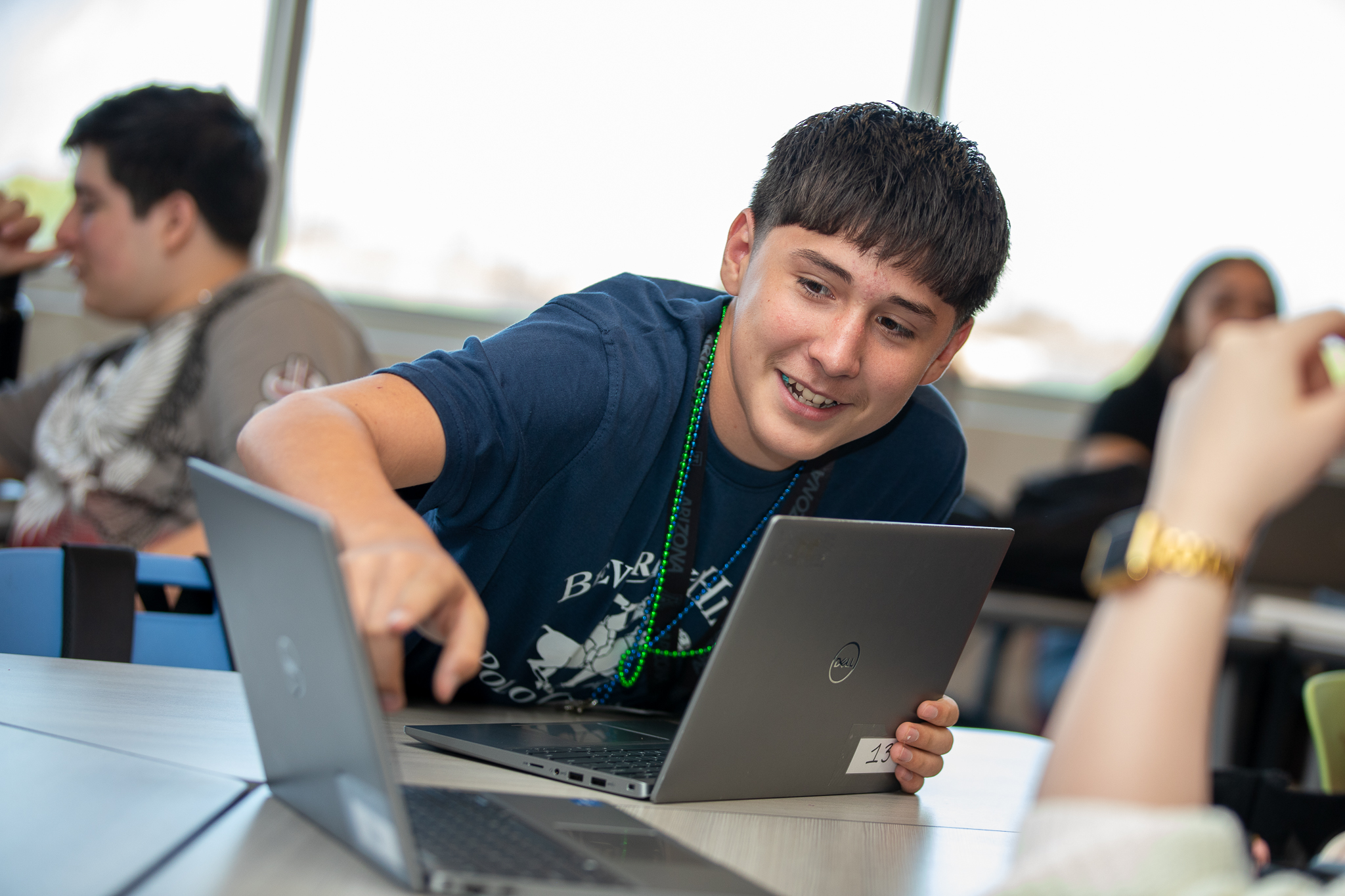A teen boy smiles as he points to something on a classmate's laptop